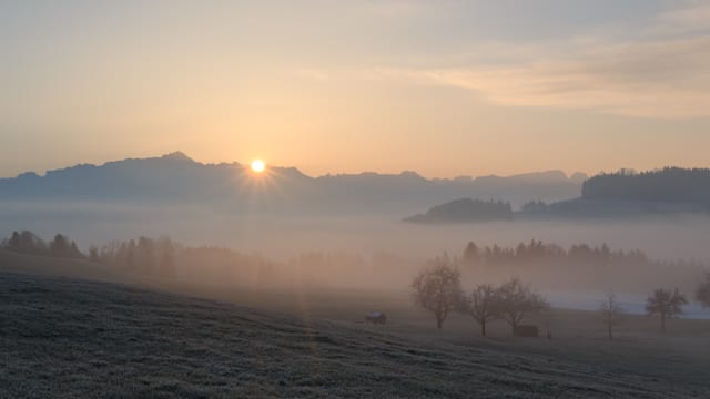 Die Sonne geht hinter den Bergen auf, über dem Tal liegen flache Nebelfelder. Die Sonne geht hinter den Bergen auf, über dem Tal liegen flache Nebelfelder.