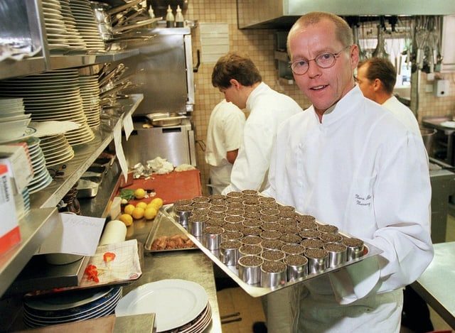Dine Mann mit kurzen roten Haaren und Brille steht in einer Küche und hält ein Tablett in der Hand. Darauf silberne kleine Schalen gefüllt mit einer braunen Masse. Dine Mann mit kurzen roten Haaren und Brille steht in einer Küche und hält ein Tablett in der Hand. Darauf silberne kleine Schalen gefüllt mit einer braunen Masse.