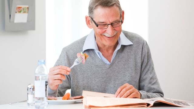 Ein älterer Mann liest Zeitung und lächelt, In der rechten Hand hält er eine Gabel mit Essen. Ein älterer Mann liest Zeitung und lächelt, In der rechten Hand hält er eine Gabel mit Essen.