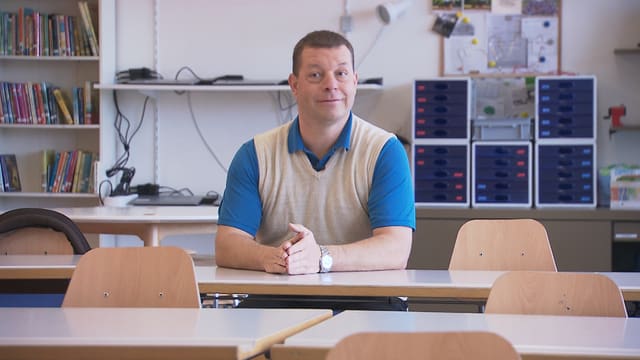 Ein Mann mittleren Alters sitzt an einem Tisch in einem Schulzimmer. Hinter ihm steht ein Bücherregal und mehrere Utensilien für den Schulgebrauch. Ein Mann mittleren Alters sitzt an einem Tisch in einem Schulzimmer. Hinter ihm steht ein Bücherregal und mehrere Utensilien für den Schulgebrauch.
