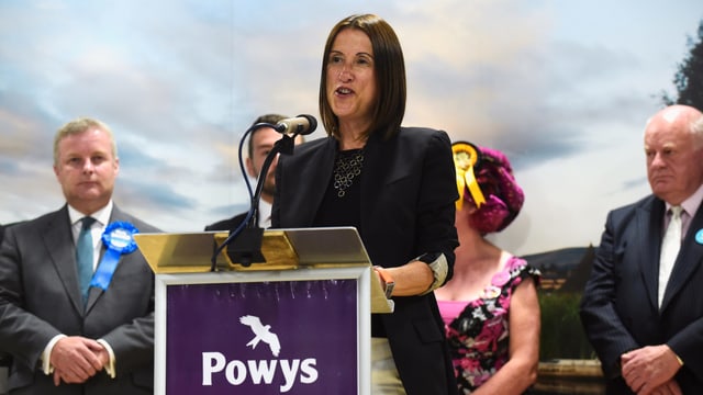 Jane Dodds an einem Rednerpult, hinter ihr vier Personen, die zuhören. Jane Dodds an einem Rednerpult, hinter ihr vier Personen, die zuhören.
