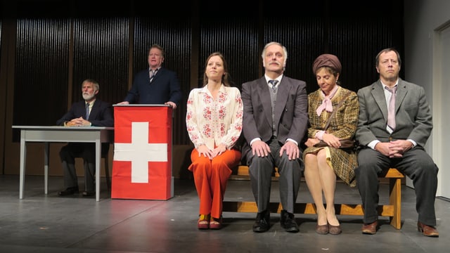 Szene in einem Landtheater: Ein Politiker steht an einem Rednerpult mit Schweizerfahne, daneben sitzen vier Leute auf einer Bank. Szene in einem Landtheater: Ein Politiker steht an einem Rednerpult mit Schweizerfahne, daneben sitzen vier Leute auf einer Bank.