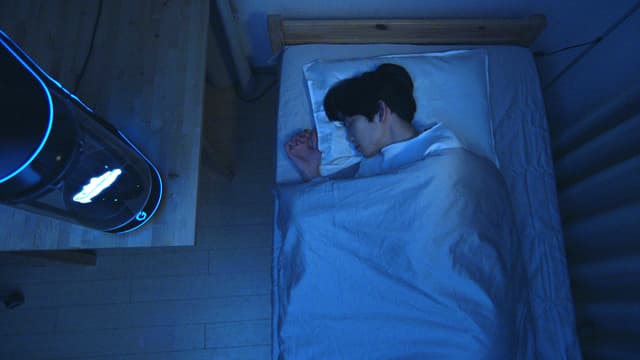 Ein junger Mann schläft auf seinem Bett, neben ihm ist ein Hologramm unter einer Glaskuppel Ein junger Mann schläft auf seinem Bett, neben ihm ist ein Hologramm unter einer Glaskuppel