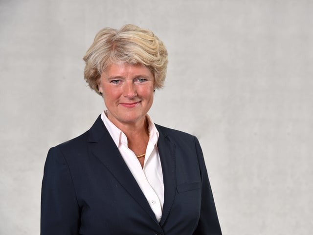 Eine Frau mit blonden, eher kürzeren haaren lächelt in die Kamera. Sie steht vor einer grauen Betonwand und trägt eine weisse Bluse und einen schwarzen Blazer. Eine Frau mit blonden, eher kürzeren haaren lächelt in die Kamera. Sie steht vor einer grauen Betonwand und trägt eine weisse Bluse und einen schwarzen Blazer.