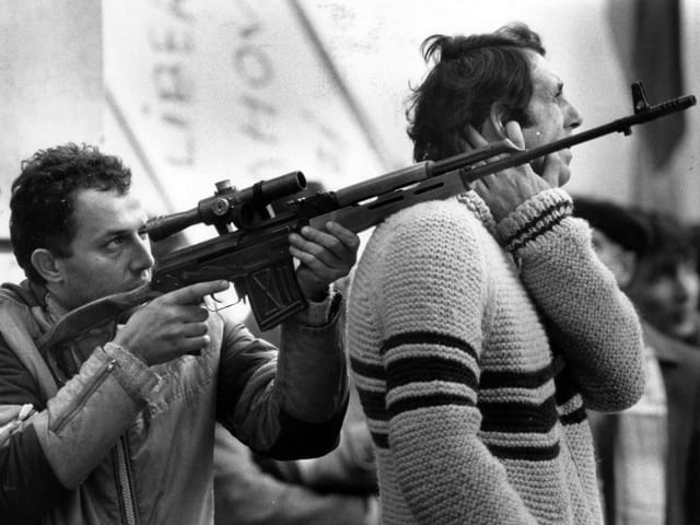 Mann mit einem Scharfschützengewehr zielend, der Gewehrlauf ruht auf der Schulter eines Zivilisten.