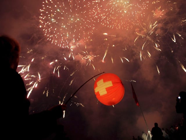 Links der Schatten eines Jungen mit Lampion in der Hand in rot mit Schweizerkreuz. Der Himmel ist voller heller Funken von Feuerwerk. Sonst ist das Foto dunkel. Links der Schatten eines Jungen mit Lampion in der Hand in rot mit Schweizerkreuz. Der Himmel ist voller heller Funken von Feuerwerk. Sonst ist das Foto dunkel.