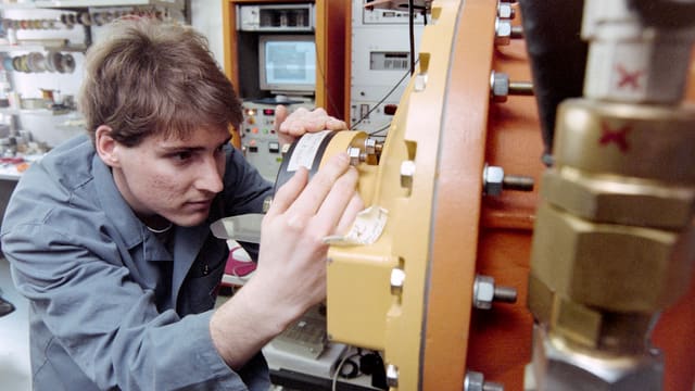 Mann in grauem Arbeitskittel schaut sich ein oranges Werkstück genau an. Mann in grauem Arbeitskittel schaut sich ein oranges Werkstück genau an.