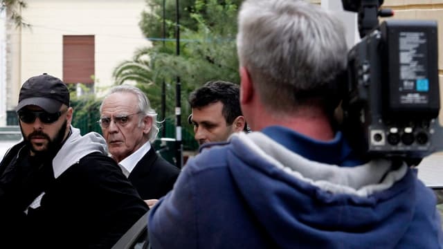 Akis Tsohatzopoulos wird abgeführt - im Vordergrund ein Kameramann, der das Ganze filmt, von hinten. Akis Tsohatzopoulos wird abgeführt - im Vordergrund ein Kameramann, der das Ganze filmt, von hinten.