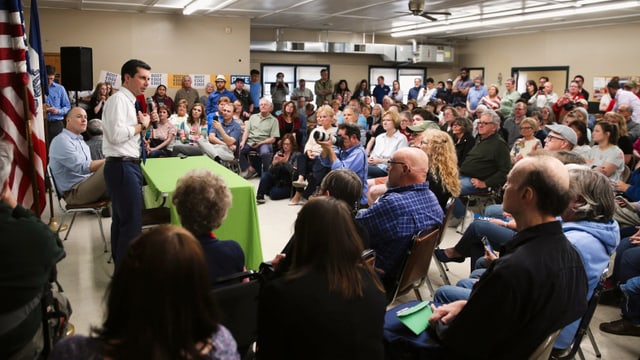 Buttigieg spricht zu Menschen in einem Saal. Buttigieg spricht zu Menschen in einem Saal.