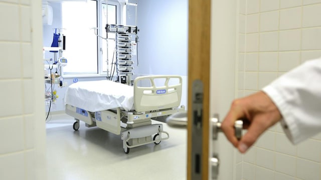 Eine Hand öffnet die Tür eines Spitalzimmers, darin ein leeres Spitalbett und diverse Geräte.