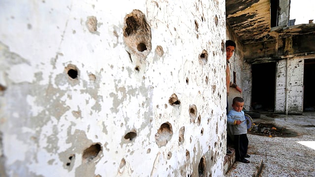 Eine Häuserwand in Aleppo in Syrien. Sie ist von Schüssen durchlöchert. An deren Ende sind ein Mann und ein Kind zu sehen, die beide aus der Ruine nach draussen spähen. Eine Häuserwand in Aleppo in Syrien. Sie ist von Schüssen durchlöchert. An deren Ende sind ein Mann und ein Kind zu sehen, die beide aus der Ruine nach draussen spähen.