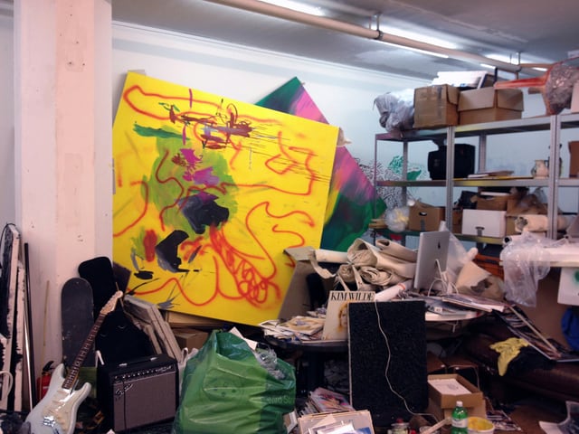 Blick von der Raummitte an eine Wand, wo Bilder übereinander aufgestellt sind. Daneben ein Regal mit verschiedenen Schachteln, davor ein Pult mit Bildschirm. Darauf und davor stapeln sich allerlei Dinge wie Leinwände, Platten, E-Gitarre und Verstärker. Blick von der Raummitte an eine Wand, wo Bilder übereinander aufgestellt sind. Daneben ein Regal mit verschiedenen Schachteln, davor ein Pult mit Bildschirm. Darauf und davor stapeln sich allerlei Dinge wie Leinwände, Platten, E-Gitarre und Verstärker.