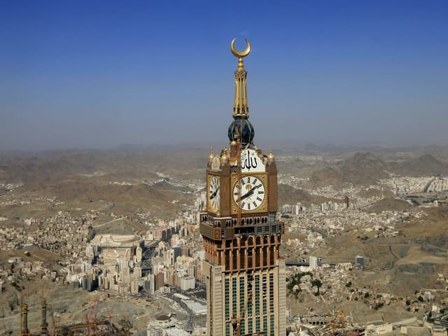 Ein riesiger Turm mit Turmuhr und goldener Mondsichel steht unter blauem Himmel inmiten einer Stadt, die in der Wüste steht. Ein riesiger Turm mit Turmuhr und goldener Mondsichel steht unter blauem Himmel inmiten einer Stadt, die in der Wüste steht.