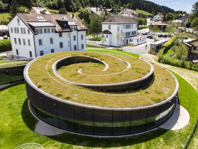 Ein schneckenförmiges Gebäude mit einer Wiese und Häusern drumherum Ein schneckenförmiges Gebäude mit einer Wiese und Häusern drumherum