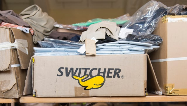 Offene Kartonschachtel mit der Aufschrift "Switcher" und Kleidern in Plastikverpackungen, die oben herausschauen. Offene Kartonschachtel mit der Aufschrift "Switcher" und Kleidern in Plastikverpackungen, die oben herausschauen.