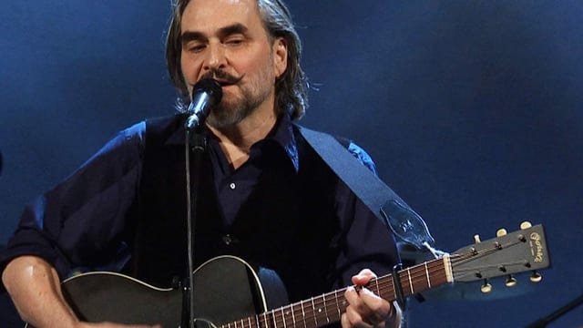 Mann mit auffälligem Schnauz spielt auf der Bühne Gitarre und singt ins Mikrofon. Mann mit auffälligem Schnauz spielt auf der Bühne Gitarre und singt ins Mikrofon.