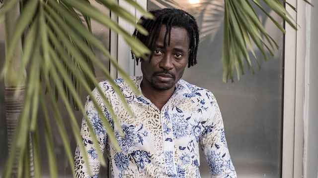 Bisi Alimi trägt ein blau-weisses Hemd. Er steht hinter einem Palmwedel. Bisi Alimi trägt ein blau-weisses Hemd. Er steht hinter einem Palmwedel.