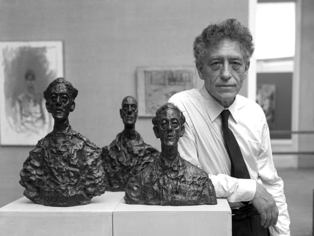 Alberto Giacometti, mit Hemd und Krawatte, an einen Sockel gelehnt, auf dem drei Büsten stehen.