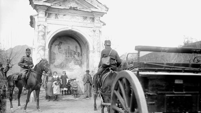 Soldaten auf Pferden gehen an einer alten Kapelle vorbei, vor der Zivilisten stehen.
