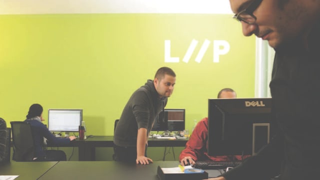 LIIP-Mitarbeitende sitzen und stehen an PCs vor einer grün gestrichenen Wand mit dem Firmenlogo.