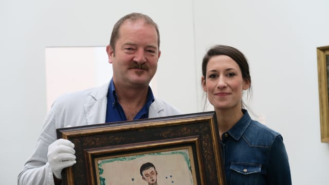 Ein Mann mit weissen handschuhen hält ein Gemälde.Rechts von ihm steht eine junge Frau. Ein Mann mit weissen handschuhen hält ein Gemälde.Rechts von ihm steht eine junge Frau.