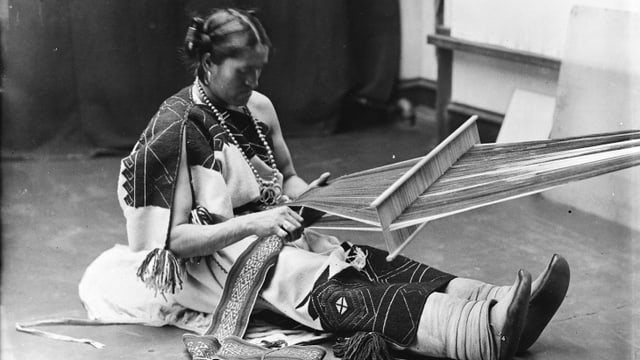 Schwarzweiss Foto eines indigenen Mannes, der Frauenkleidung trägt und webt. Schwarzweiss Foto eines indigenen Mannes, der Frauenkleidung trägt und webt.