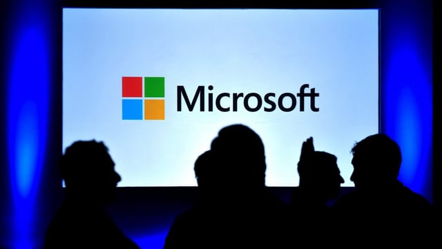 Microsoft-Logo auf einem Bildschirm, einige Menschen davor. Microsoft-Logo auf einem Bildschirm, einige Menschen davor.