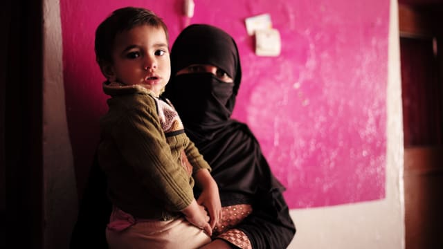 Schwarz verhüllte Frau mit Kind auf dem Arm vor rosa Wand. Schwarz verhüllte Frau mit Kind auf dem Arm vor rosa Wand.