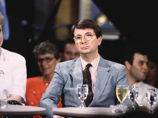 Mann mit Brille und Anzug im Stil der 1990er-Jahren sitzt an einem Tisch. Vor ihm ein Wasserglas. Mann mit Brille und Anzug im Stil der 1990er-Jahren sitzt an einem Tisch. Vor ihm ein Wasserglas.