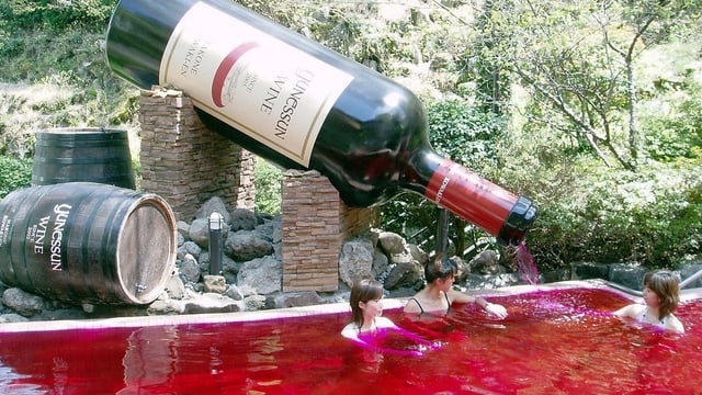 In einem Bad mit rotem Wasser sitzen drei junge Frauen. Hinter dem Bad ist eine riesige Weinflasche installiert, aus der das rote Wasser direkt ins Bad fliesst. In einem Bad mit rotem Wasser sitzen drei junge Frauen. Hinter dem Bad ist eine riesige Weinflasche installiert, aus der das rote Wasser direkt ins Bad fliesst.