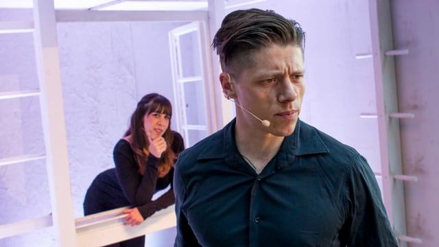 Theater: Ein Mann mit Mikrophon, hinter ihm lehnt eine Frau im Fenster. Theater: Ein Mann mit Mikrophon, hinter ihm lehnt eine Frau im Fenster.