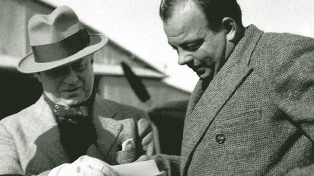 Aufnahme von Saint-Exupéry und Marcel Peyrouton, die gemeinsam in Unterlagen schauen. Im Hintergrund ist unscharf ein Flugzeug zu sehen. Aufnahme von Saint-Exupéry und Marcel Peyrouton, die gemeinsam in Unterlagen schauen. Im Hintergrund ist unscharf ein Flugzeug zu sehen.