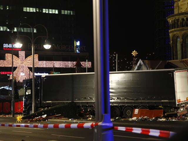 Unter einem grossen, schwarzen LKW hinter Polizeiabsperrungen schauen umgerissene Weihnachtsbäume hervor. Unter einem grossen, schwarzen LKW hinter Polizeiabsperrungen schauen umgerissene Weihnachtsbäume hervor.