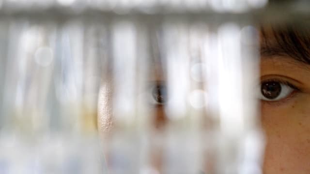 Nahaufnahme eines Gesichts. Erkennbar sind nur die Augen, im Vordergrund sind unscharf Reagenzgläser zu sehen. Nahaufnahme eines Gesichts. Erkennbar sind nur die Augen, im Vordergrund sind unscharf Reagenzgläser zu sehen.