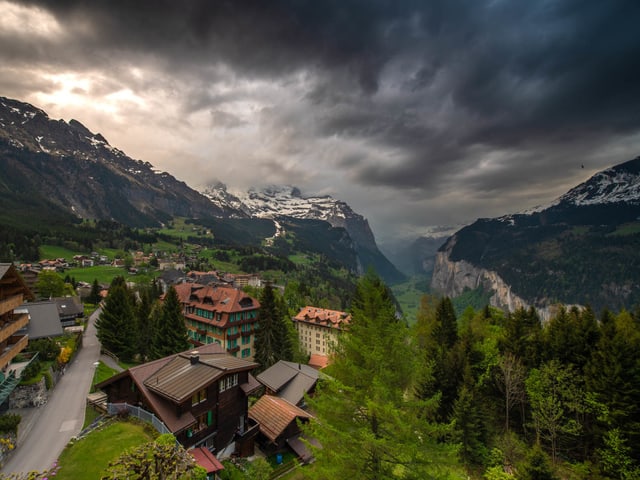 Bergwelt mit dunklen Wolken am Himmel und einem schroffen Tal. Im Vordergrund stehen Häuser ein Strassen führt hindurch.