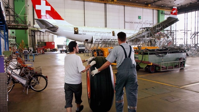 Zwei Mitarbeiter rollen einen fast manhshohen Flugzeugreifen zu einem im Hintergrund in der Halle stehenden Flugzeug mit dem Schweizerkreuz an der Heckflosse. Zwei Mitarbeiter rollen einen fast manhshohen Flugzeugreifen zu einem im Hintergrund in der Halle stehenden Flugzeug mit dem Schweizerkreuz an der Heckflosse.