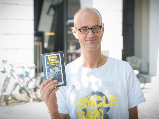 Mann im T-Shirt in einer Altstadt-Gasse, der lachend ein Buch hochhält.
