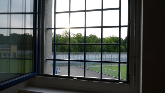 Blick durch ein vergittertes Fenster. Zu sehen ist ein Fussballplatz, dahinter eine Mauer und ein Wald. Die Sonne scheint. Blick durch ein vergittertes Fenster. Zu sehen ist ein Fussballplatz, dahinter eine Mauer und ein Wald. Die Sonne scheint.