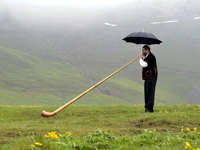 Ein Alphornblässer hält in einer Hand das Instrument und in der anderen Hand einen Regenschirm Ein Alphornblässer hält in einer Hand das Instrument und in der anderen Hand einen Regenschirm
