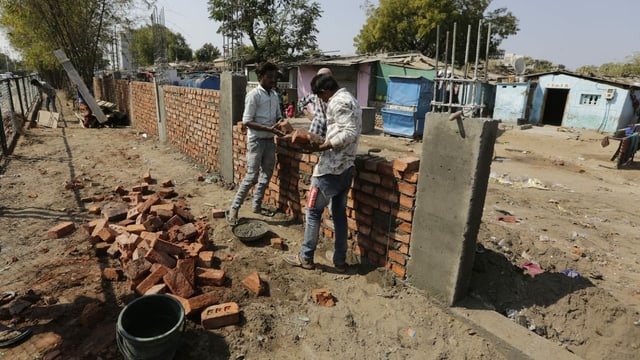 Zwei Arbeiter bauen eine Mauer aus Ziegelsteinen. Im Hintergrund ein Slum. Zwei Arbeiter bauen eine Mauer aus Ziegelsteinen. Im Hintergrund ein Slum.