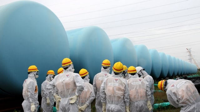 Arbeiter in Schutzanzügen stehen vor grossen, blauen, runden Containern Arbeiter in Schutzanzügen stehen vor grossen, blauen, runden Containern
