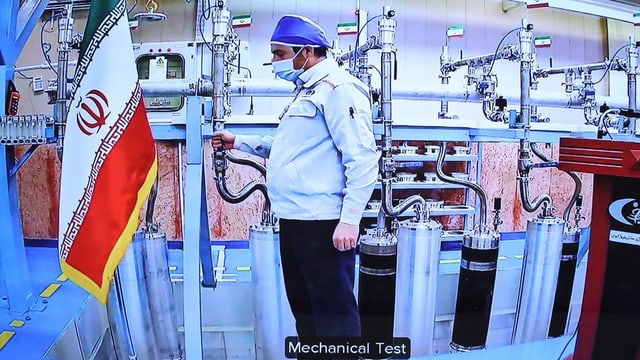 Ein Mann mit Haube steht in einem Raum mit Behältern und Rohen aus Metall. Links im Bild hängt eine iranische Flagge. Ein Mann mit Haube steht in einem Raum mit Behältern und Rohen aus Metall. Links im Bild hängt eine iranische Flagge.