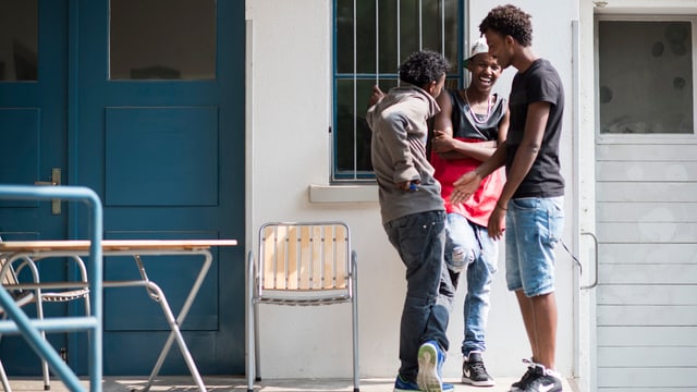 Drei jugendliche Flüchtlinge stehen vor einem weissen Gebäude mit blauer Tür Drei jugendliche Flüchtlinge stehen vor einem weissen Gebäude mit blauer Tür