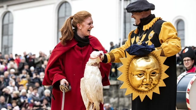 Eine Frau im roten Mantel hält eine tote Gans, daneben ein kostümierter Mann mit Sonnenmaske. Eine Frau im roten Mantel hält eine tote Gans, daneben ein kostümierter Mann mit Sonnenmaske.