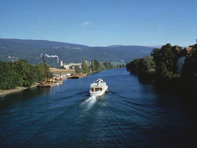Ein Kursschiff fährt durch den mit Bäumen gesäumten Kanal. Im Hintergrund die Jurahöhen und blauer Himmel. Ein Kursschiff fährt durch den mit Bäumen gesäumten Kanal. Im Hintergrund die Jurahöhen und blauer Himmel.