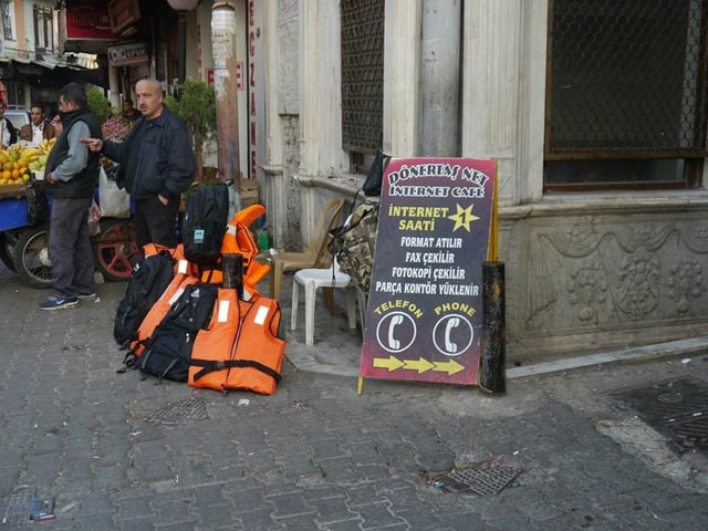Ecke einer Gasse, Schwimmwesten am Boden, Verkäufer, ein Schild Ecke einer Gasse, Schwimmwesten am Boden, Verkäufer, ein Schild