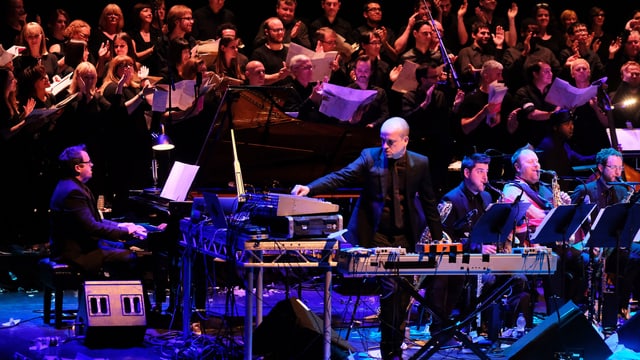 Ein Mann steht am Mischpult auf einer Bühne, eine Big Band ist im Hintergrund. Ein Mann steht am Mischpult auf einer Bühne, eine Big Band ist im Hintergrund.