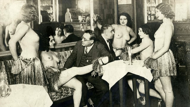 Männer in Anzug und nackte Frauen sitzen und stehen in einer Gaststube um einen Tisch.