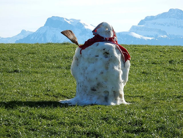 Leicht angeschmolzener Schneemann auf grüner Wiesi, im HIntergrund verschneite Berge. Leicht angeschmolzener Schneemann auf grüner Wiesi, im HIntergrund verschneite Berge.