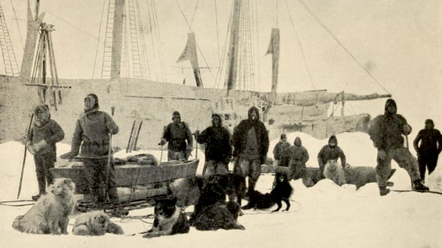 Altes Foto: Männer in Winterkleidung im Schnee neben Hundeschlitten. Im Hintergrund ein grosses Segelschiff. Altes Foto: Männer in Winterkleidung im Schnee neben Hundeschlitten. Im Hintergrund ein grosses Segelschiff.